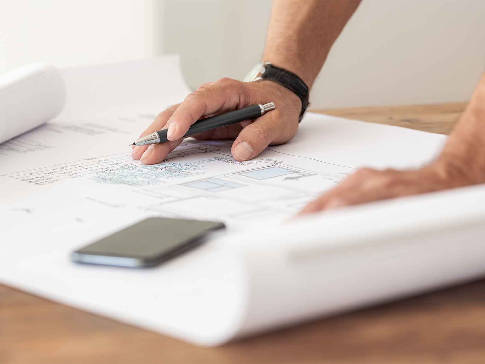 Zwei Hände stützen sich auf einen Bauplan. Der Mann hält in der rechten Hand einen Stift, vor ihm liegt ein Mobiltelefon.