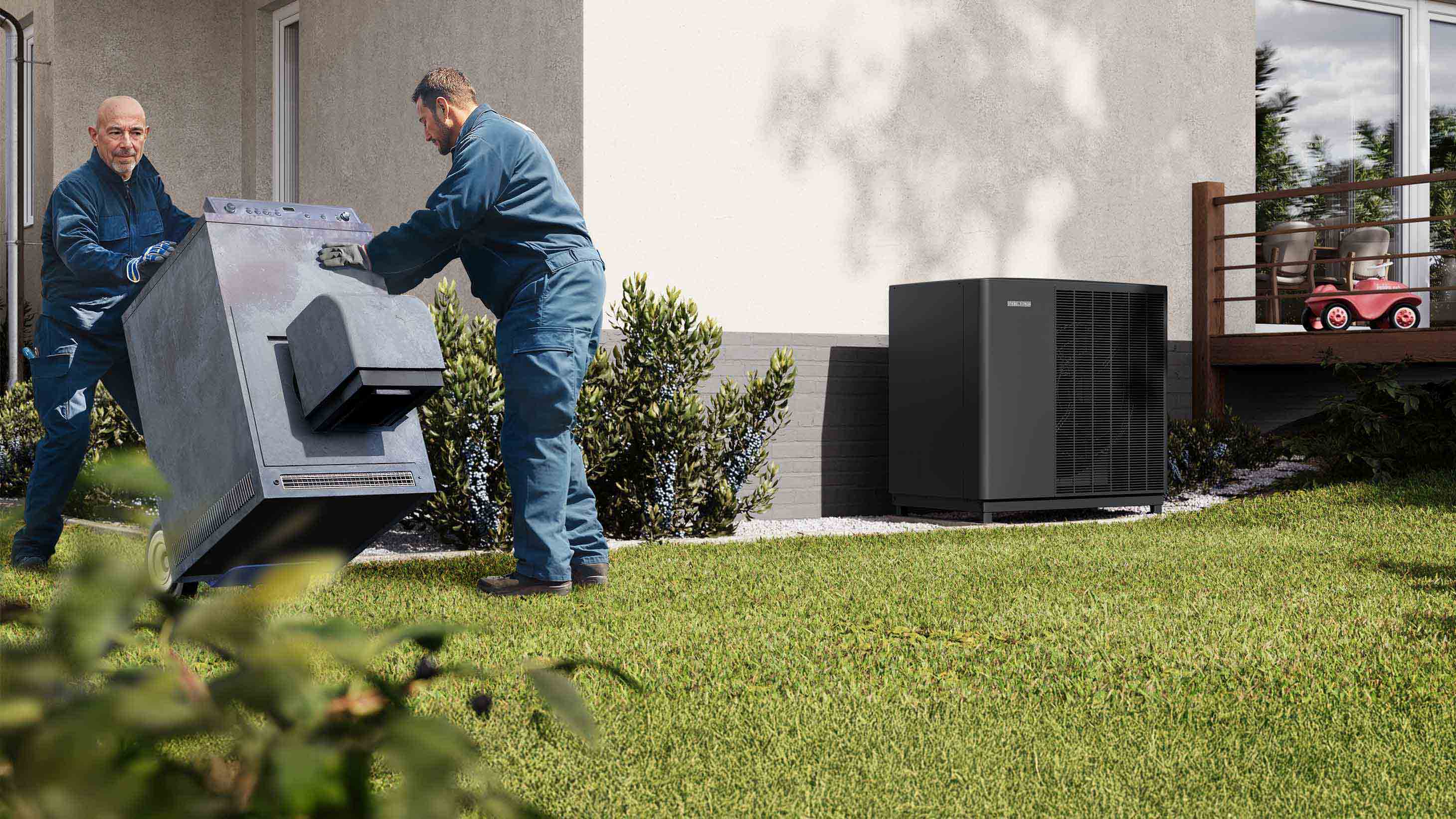 Zwei Handwerker transportieren eine alte Ölheizung aus einem Einfamilienhaus. An der Fassade steht eine neue Wärmepumpe.