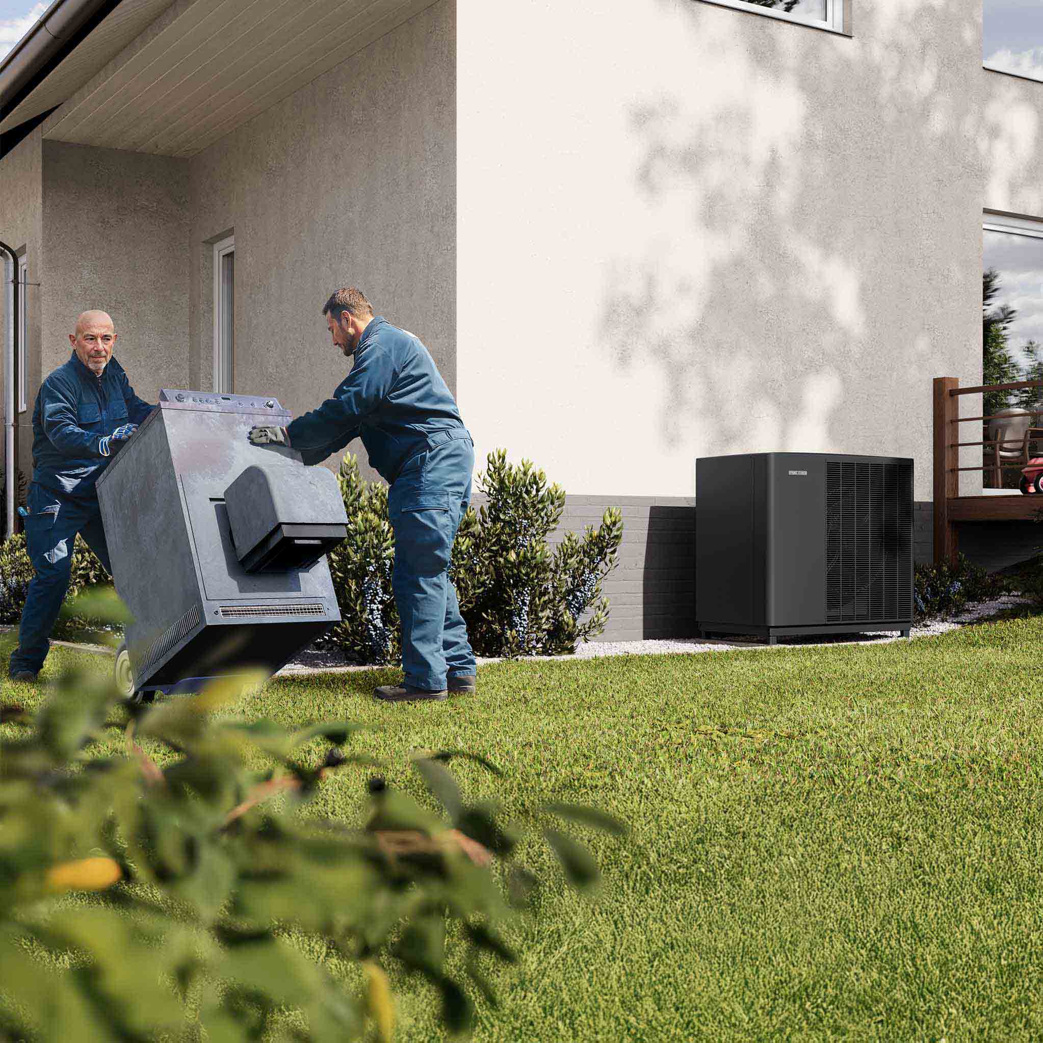 Zwei Installateure tragen eine Ölheizung aus dem Haus, vor dem Haus steht eine STIEBEL ELTRON-Luft Wasser Wärmepumpe