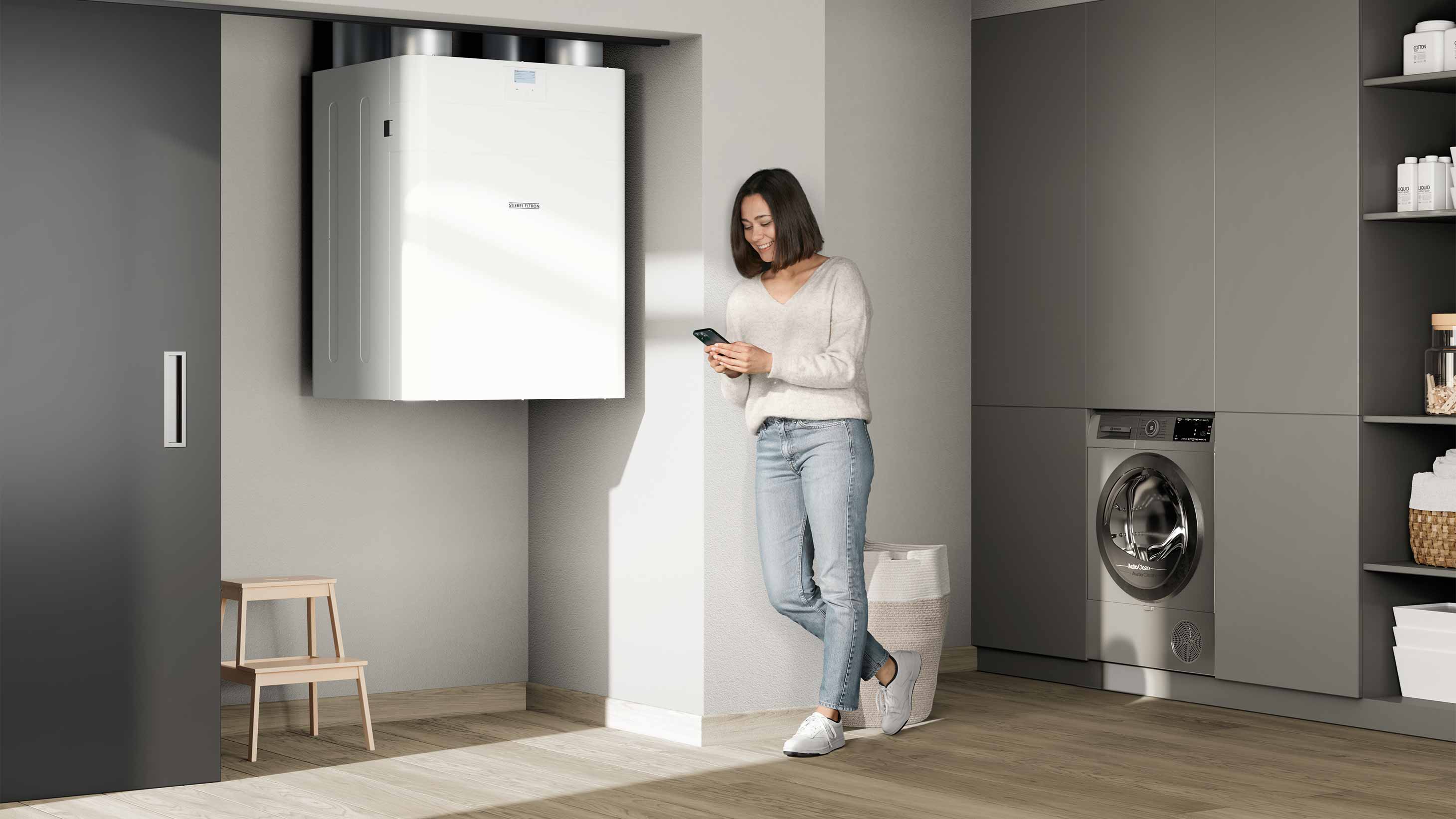 Eine Frau lehnt an der Wand und schaut auf ihr Handy. Neben ihr hängt ein zentrales Lüftungsgerät an der Wand.