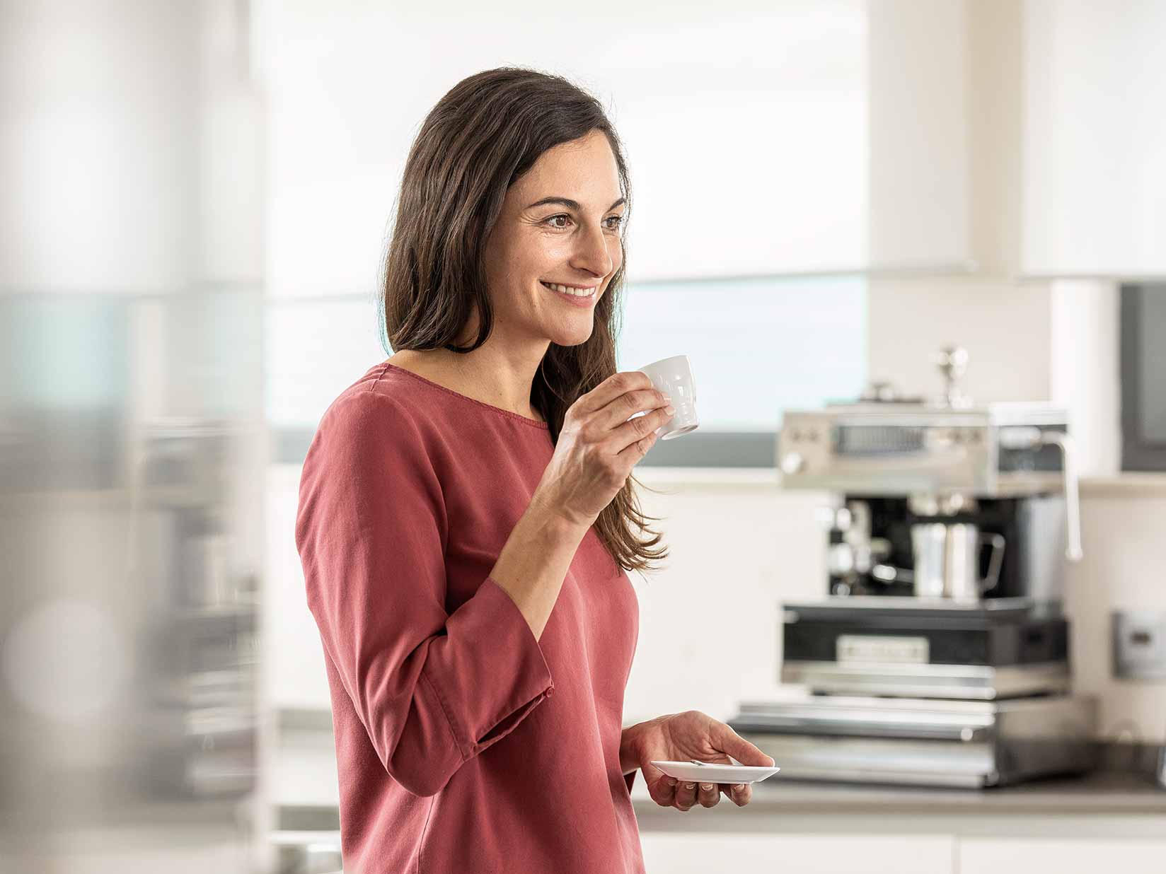 Frau hält eine Espressotasse in der Hand. Im Hintergrund befindet sich eine Kaffeemaschine.