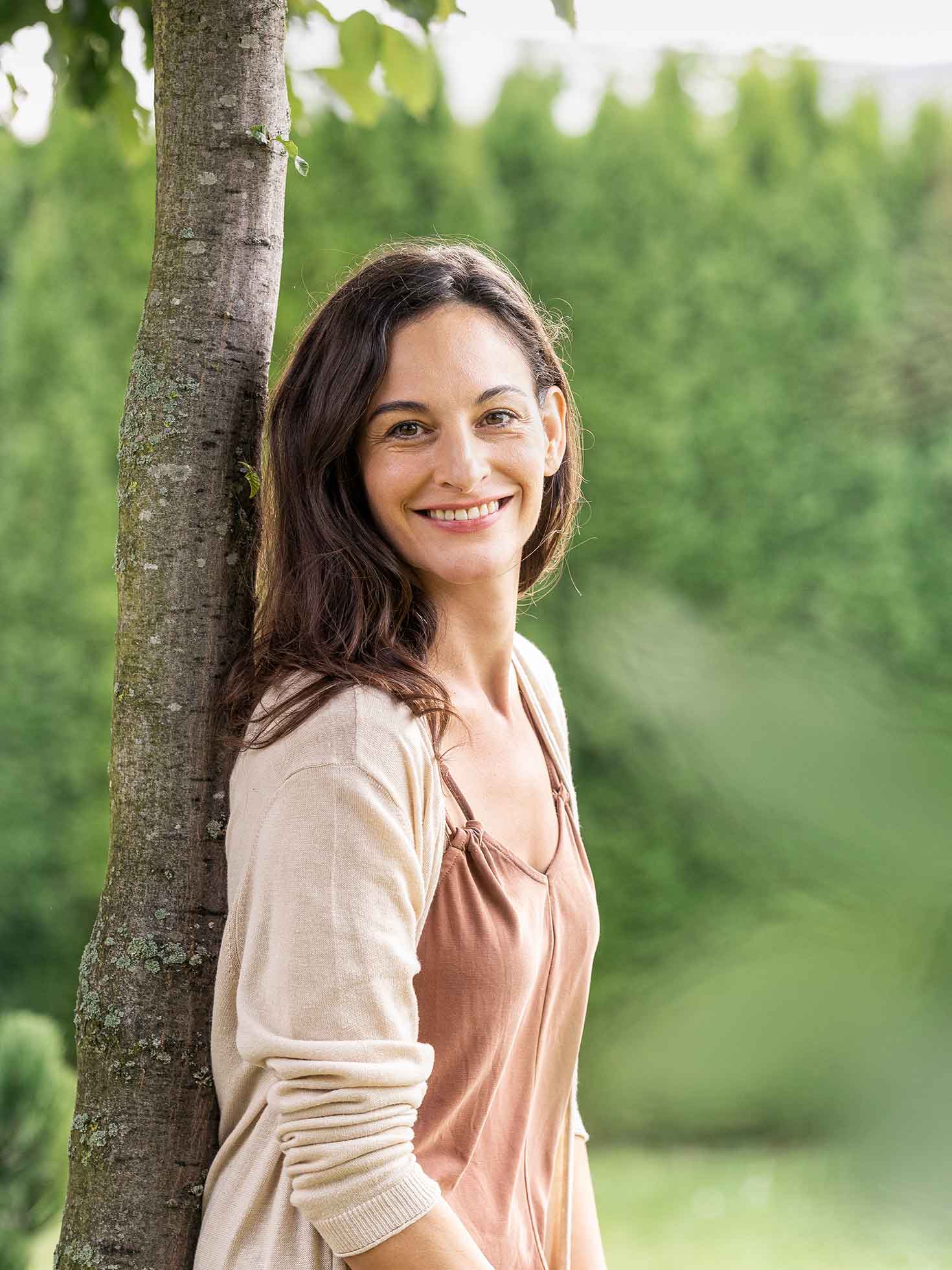 Eine lächelnde Frau lehnt sich an einem Baum. Die Frau steht vor einer grünen Hecke, welche verschwommen dargestellt ist.