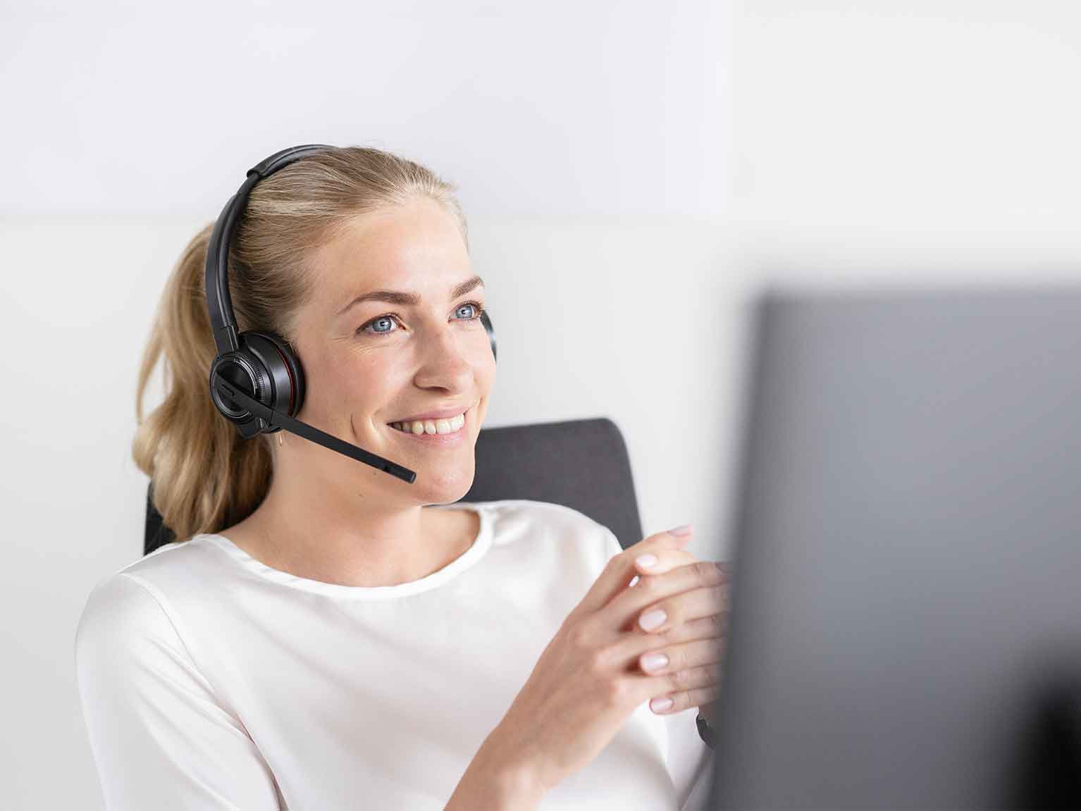 Eine lächelnde Frau sitzt auf einem Stuhl, hat die Finger ineinander verschränkt und trägt ein Headset auf dem Kopf. Vor der Frau befindet sich ein Computerbildschirm.