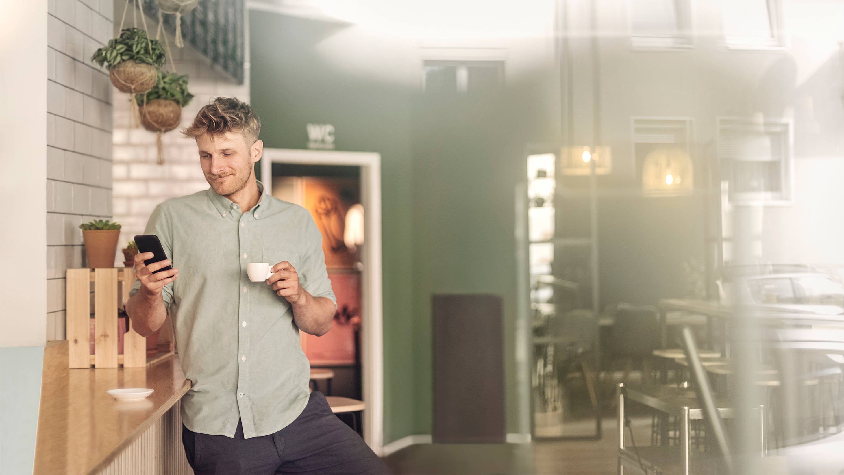 Ein Mann steht mit seinem Smartphone in der Hand an die Küchzeile gelehnt und trinkt Espresso