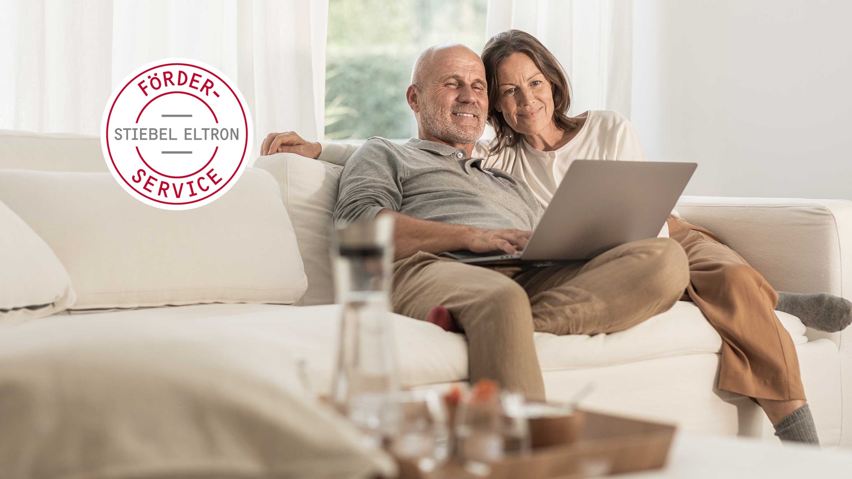 Ein Paar sitzt auf einer hellen Couch und schaut lächelnd auf einen Laptop. 