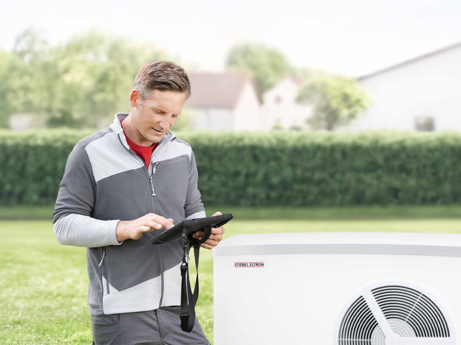 Ein Fachhandwerker steht neben einer Wärmepumpe und bedient sein Tablet