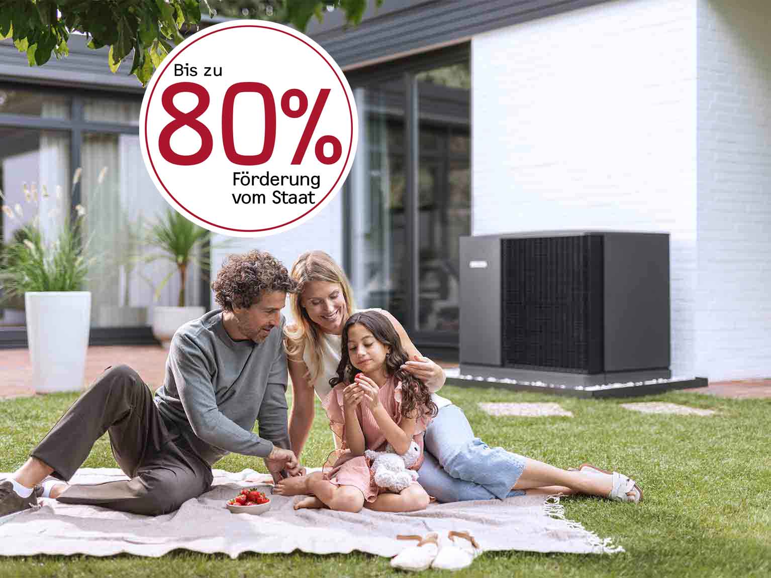 Familie picknickt im Garten, im Hintergrund steht eine Wärmepumpe vor dem Haus