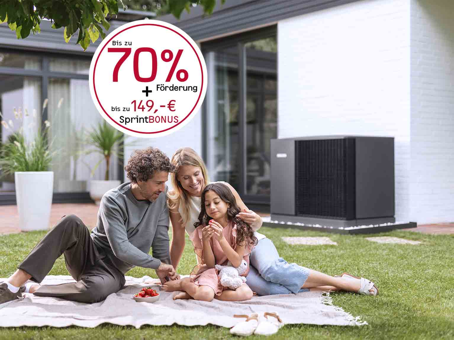 Familie picknickt im Garten, im Hintergrund steht eine Wärmepumpe vor dem Haus