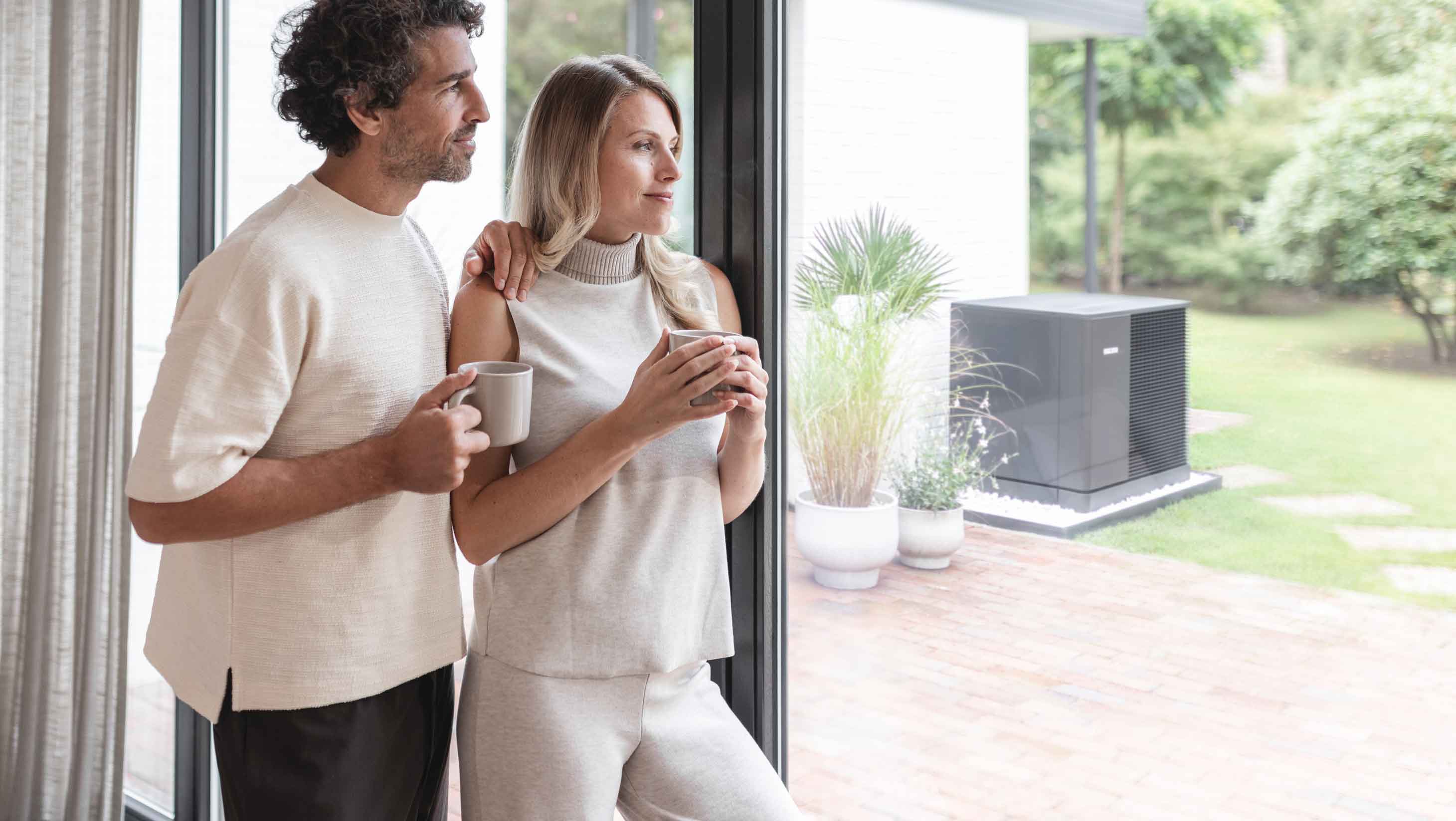 Paar steht mit Kaffeetassen in der Hand vor dem Fenster und blick auf die Wärmepumpe, die im Garten steht