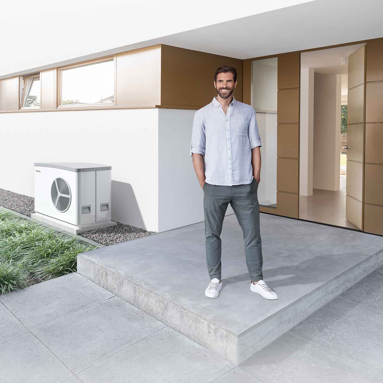 Ein Mann steht mit Händen in den Hosentaschen vor dem Eingang eines modernen Hauses. Neben dem Eingang steht eine Wärmepumpe.