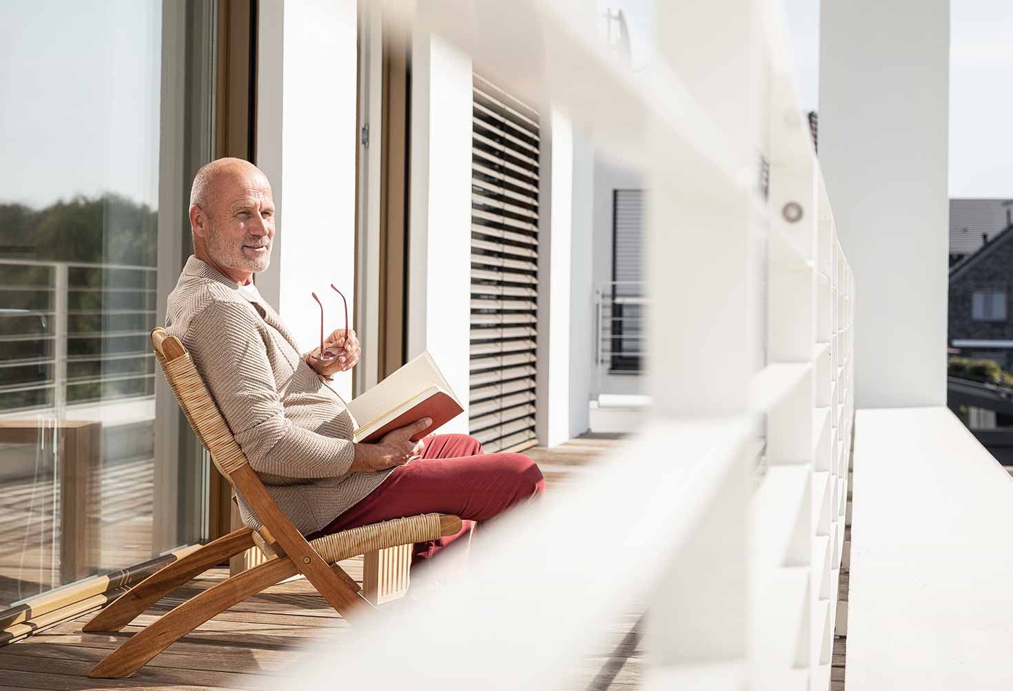 Ein Mann sitzt auf der Terasse seines Hauses und liest ein Buch. In der Hand hält er eine Brille.