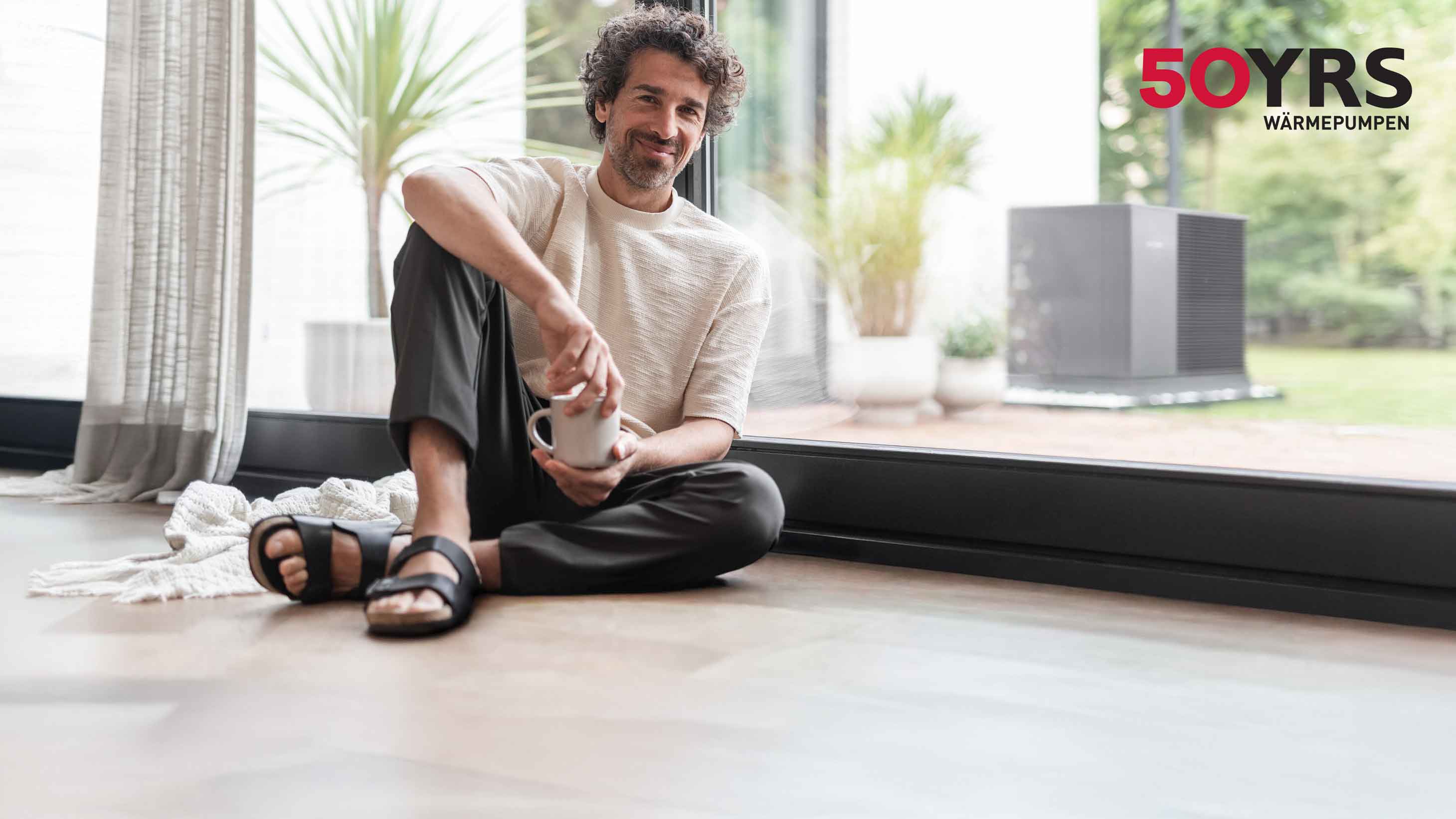 Ein Mann sitzt auf dem Boden vor einer Fensterfront und hält eine Tasse in der Hand. Im Hintergrund ist ein Garten mit einer STIEBEL ELTRON Wärmepumpe zu sehen.